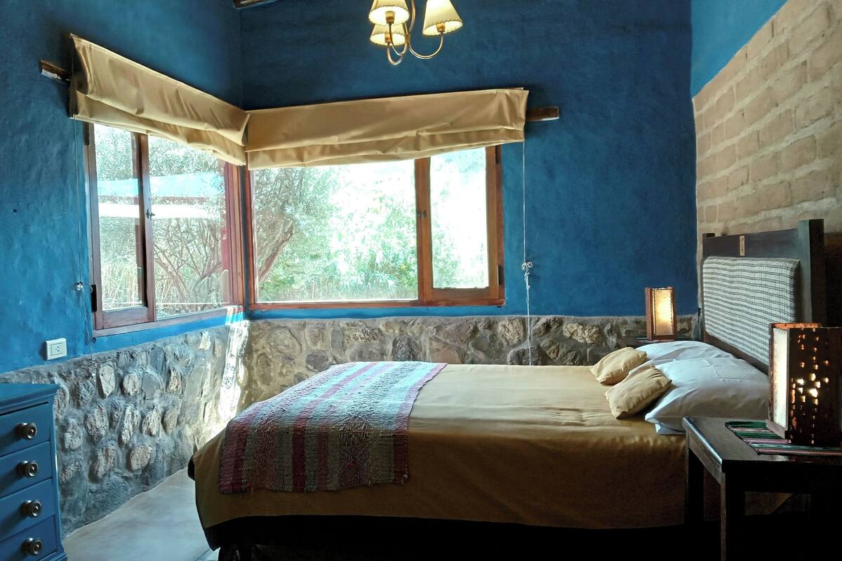 A bedroom with a bed, a dresser, a nightstand, a lamp, and a window with a view of greenery.