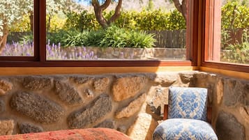 Un cuarto de spa con pared de piedra, una camilla de masaje con toallas, una silla y vista a árboles y plantas a través de amplios ventanales.