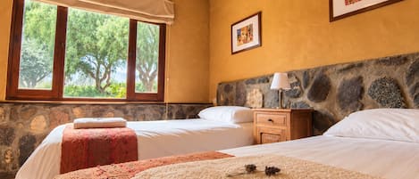 A hotel room with two beds, a stone headboard, a large window with a view of trees, and a framed picture on the wall.