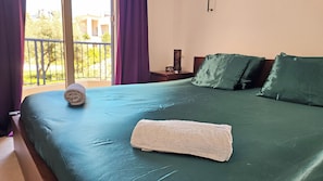 Ein Hotelzimmer mit Bett, Nachttisch, Fenster mit Blick auf Grünflächen und Gebäude sowie Balkon mit violetten Vorhängen.