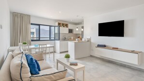 A modern living room with a sofa, a dining table, and a kitchen area with a TV mounted on the wall. - Vacation Home 'Casa Montaña Clara Famara' with Mountain View and Wi-Fi (Caleta de Famara)