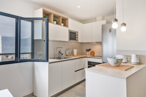 A modern kitchen with white cabinetry, a built-in microwave, a large island with a cutting board, and a view of the sea through a window. - Vacation Home 'Casa Montaña Clara Famara' with Mountain View and Wi-Fi (Caleta de Famara)