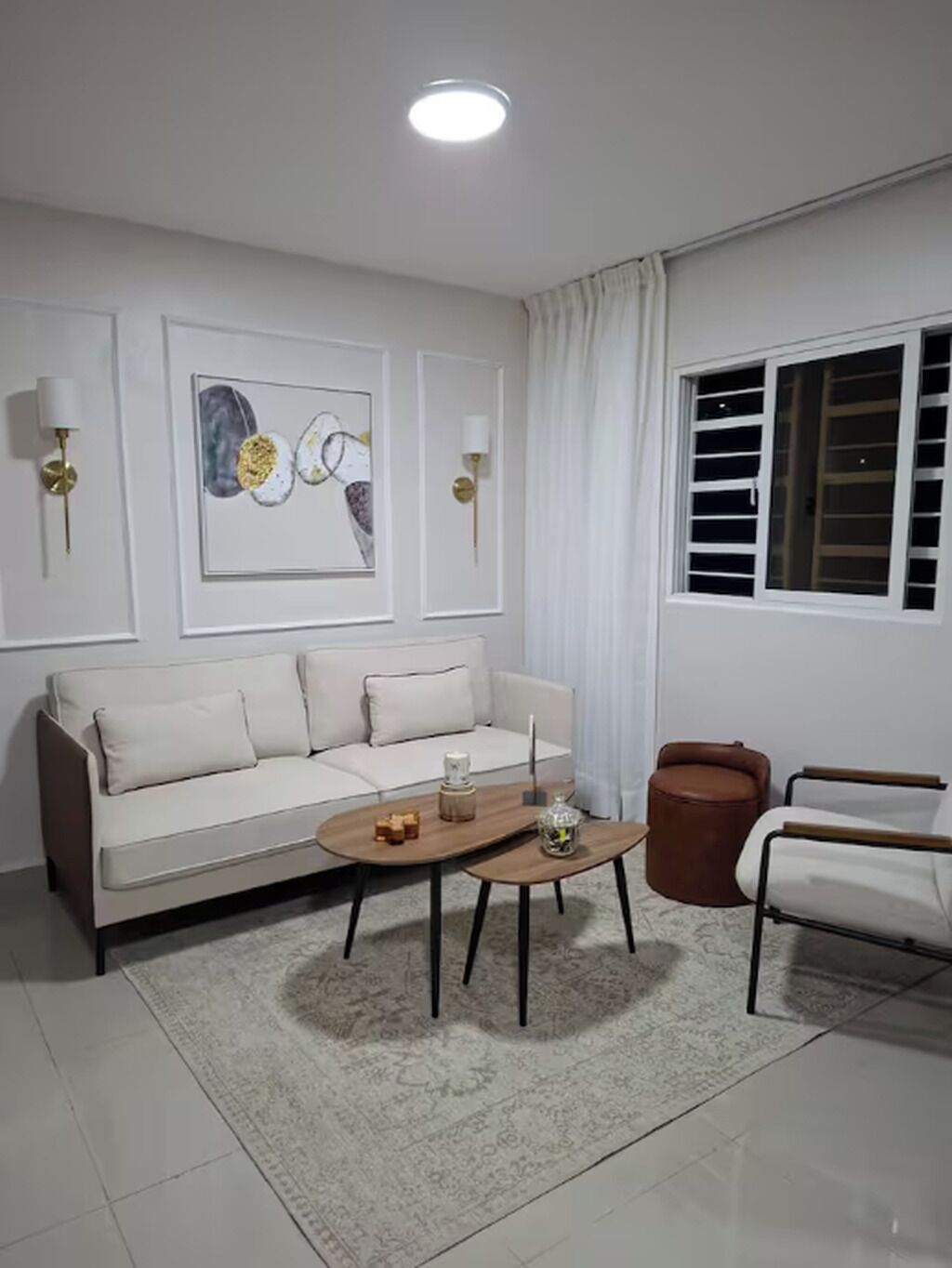 Un salon comprenant un canapé, une table basse et deux fauteuils. On y trouve une fenêtre avec des stores, un luminaire au plafond et deux lampes fixées au mur.