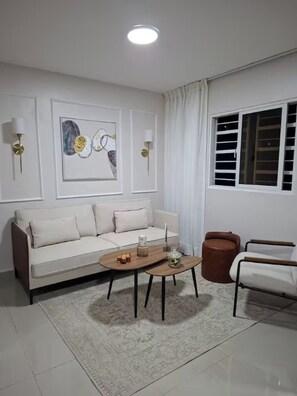 A living room with a sofa, coffee table, and two chairs. There is a window with blinds, a ceiling light, and two wall-mounted lamps. - Nice Apartment in Santo Domingo with A/C, Wi-Fi, and Parking (Santo Domingo)