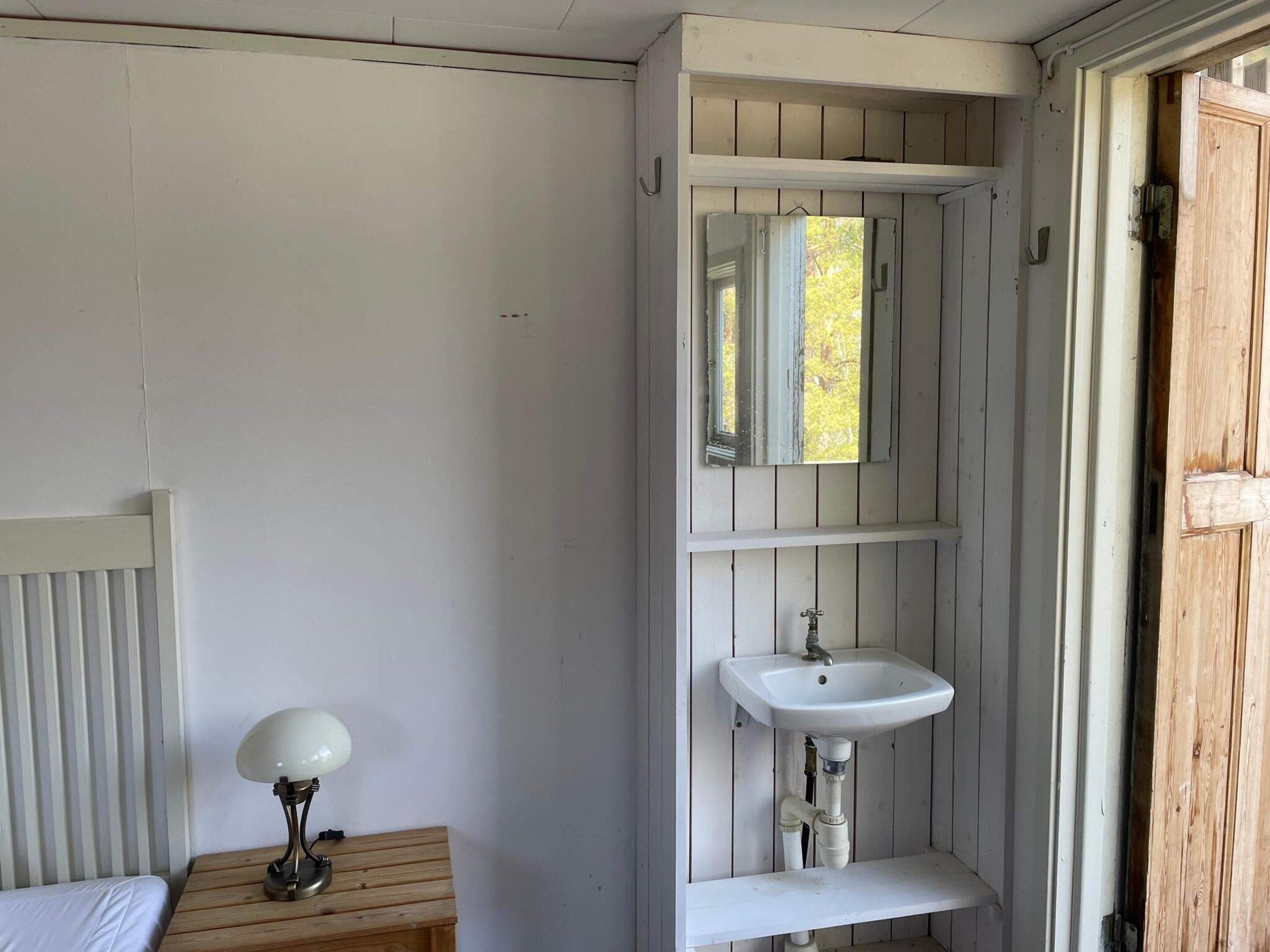 Een kleine, eenvoudige accommodatie met een houten bed, een nachtkastje met een lamp, een witte wastafel en een spiegel.