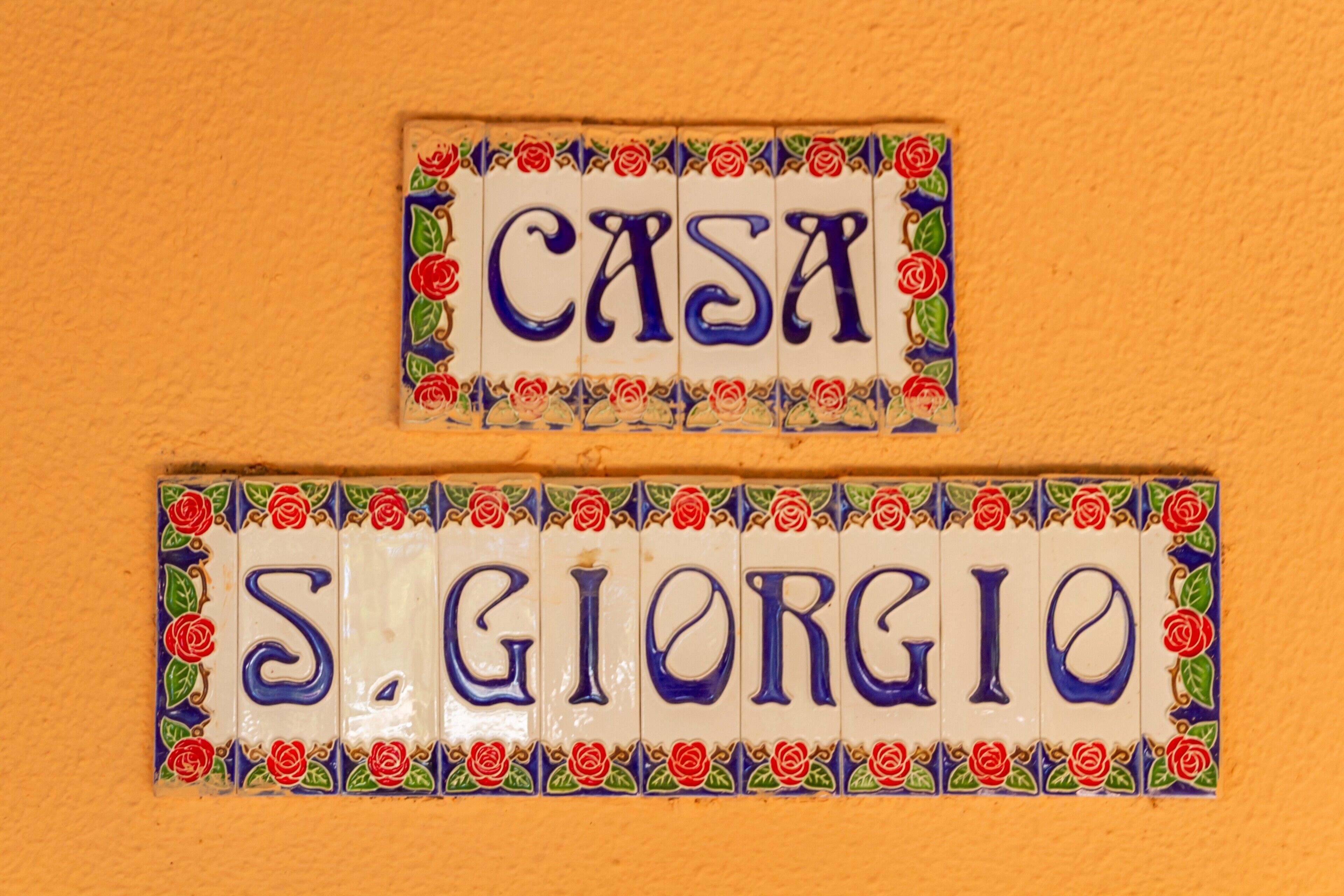 Zwei Keramikfliesen mit den Aufschriften 'CASA' und 'S. GIORGIO' an einer strukturierten orangefarbenen Wand.
