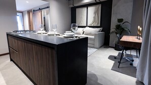 Ett modernt kök med en central köksö, en matplats med bord och stolar samt ett vardagsrum med soffa och gardiner.