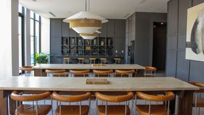 Uma área de jantar moderna com uma grande mesa retangular, várias cadeiras, um lustre pendurado e uma estante de livros ao fundo.