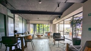 Ein geräumiges Zimmer mit Holzboden, einem Essbereich mit Stühlen und Tisch, einer Couch und einem großen Fenster mit Blick auf die grüne Umgebung.