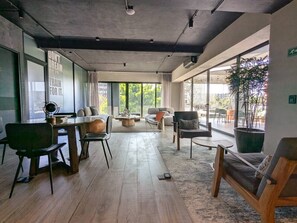 Ein geräumiges Zimmer mit Holzboden, einem Essbereich mit Stühlen und Tisch, einer Couch und einem großen Fenster mit Blick auf die grüne Umgebung.