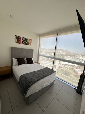 Een slaapkamer met een bed, nachtkastje, raam met uitzicht op een stadsbeeld en kunst aan de muur.
