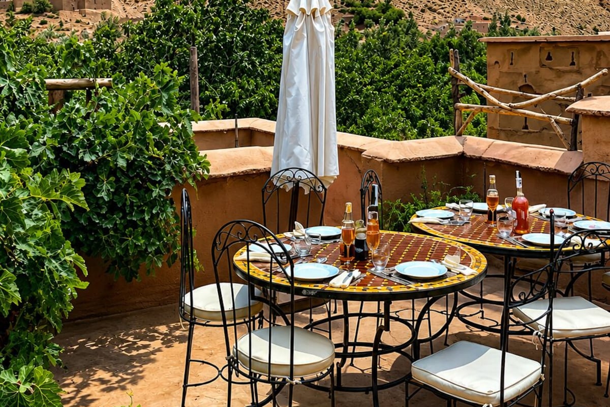 Un área de comedor al aire libre con una mesa dispuesta para cuatro personas, rodeada de sillas, en un patio con vista a un paisaje montañoso.