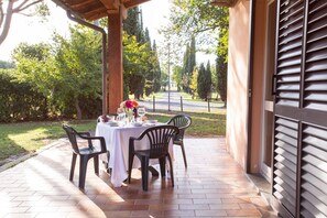 Eine Terrasse im Freien mit Esstisch und Stühlen, mit Blick auf einen von Bäumen gesäumten Weg.