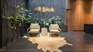 Een receptiezone met een modern design, met een houten wand waarop de naam 'ATENA' verlicht is, twee leren fauteuils, een ronde salontafel en een vachttapijt.