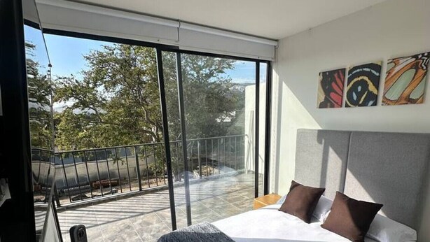 Une chambre moderne avec un grand lit, une télévision et un balcon offrant une vue sur les arbres et les bâtiments.