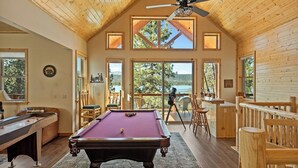 Une table de billard dans une pièce aux murs en bois et dotée d’un ventilateur de plafond, de grandes fenêtres offrant une vue sur l’extérieur.