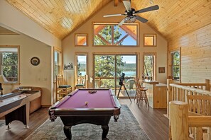 A pool table in a room with wooden walls and a ceiling fan, large windows offering a view of the outdoors. - New Lakefront Retreat w/ Hot Tub & Game Room (Fawnskin)