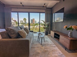 Ett modernt vardagsrum med en soffa, ett soffbord, en tv och stora fönster med utsikt över staden.