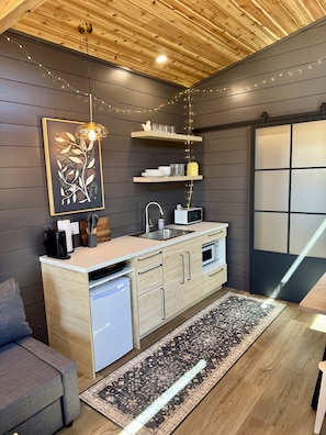 A modern kitchen with wooden cabinets, a built-in dishwasher, and a dining area with a rug. - The Birch Tiny House at Cyprus Trail (Rockbridge)