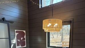 Et værelse med et træbord, to stole, en hængende lampe, et vindue med udsigt til træer, og en lyskontakt på væggen.