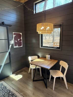 A room with a wooden table, two chairs, a hanging light fixture, a window with a view of trees, and a wall-mounted light switch.