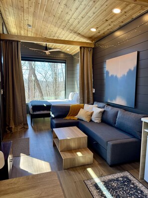 A modern interior with a wooden ceiling, a large window with curtains, a sofa with cushions, a coffee table, and a rug.