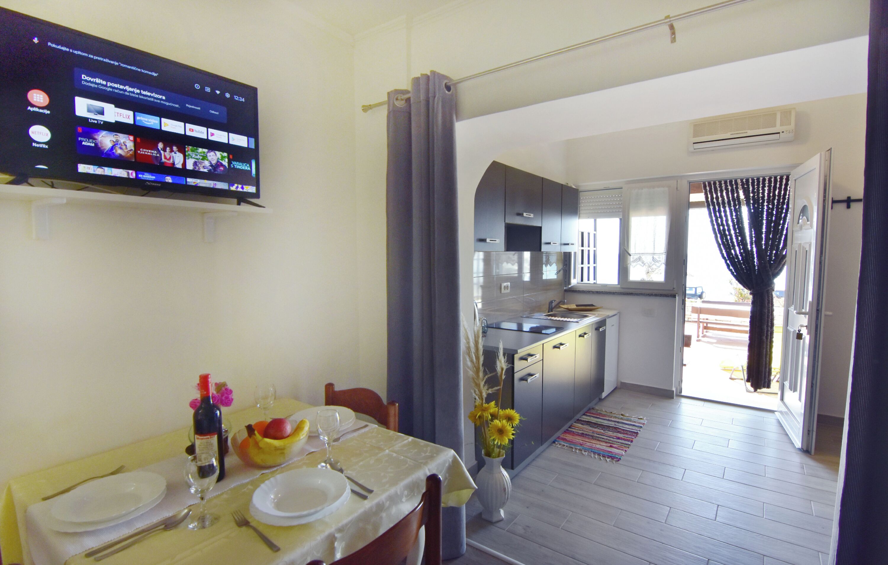 A dining area with a TV displaying a streaming service, a kitchen with a modern design, and a view through an open door.