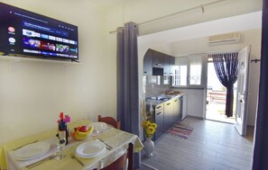 A dining area with a TV displaying a streaming service, a kitchen with a modern design, and a view through an open door. - Cozy Beachside Retreat (Umag)