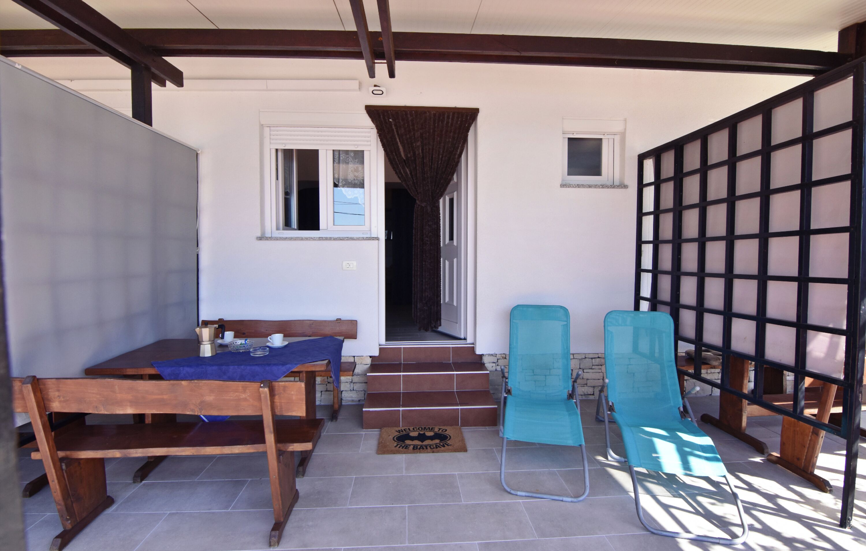 A covered patio area with a wooden table and chairs, a blue lounge chair, and a door leading to an interior space.