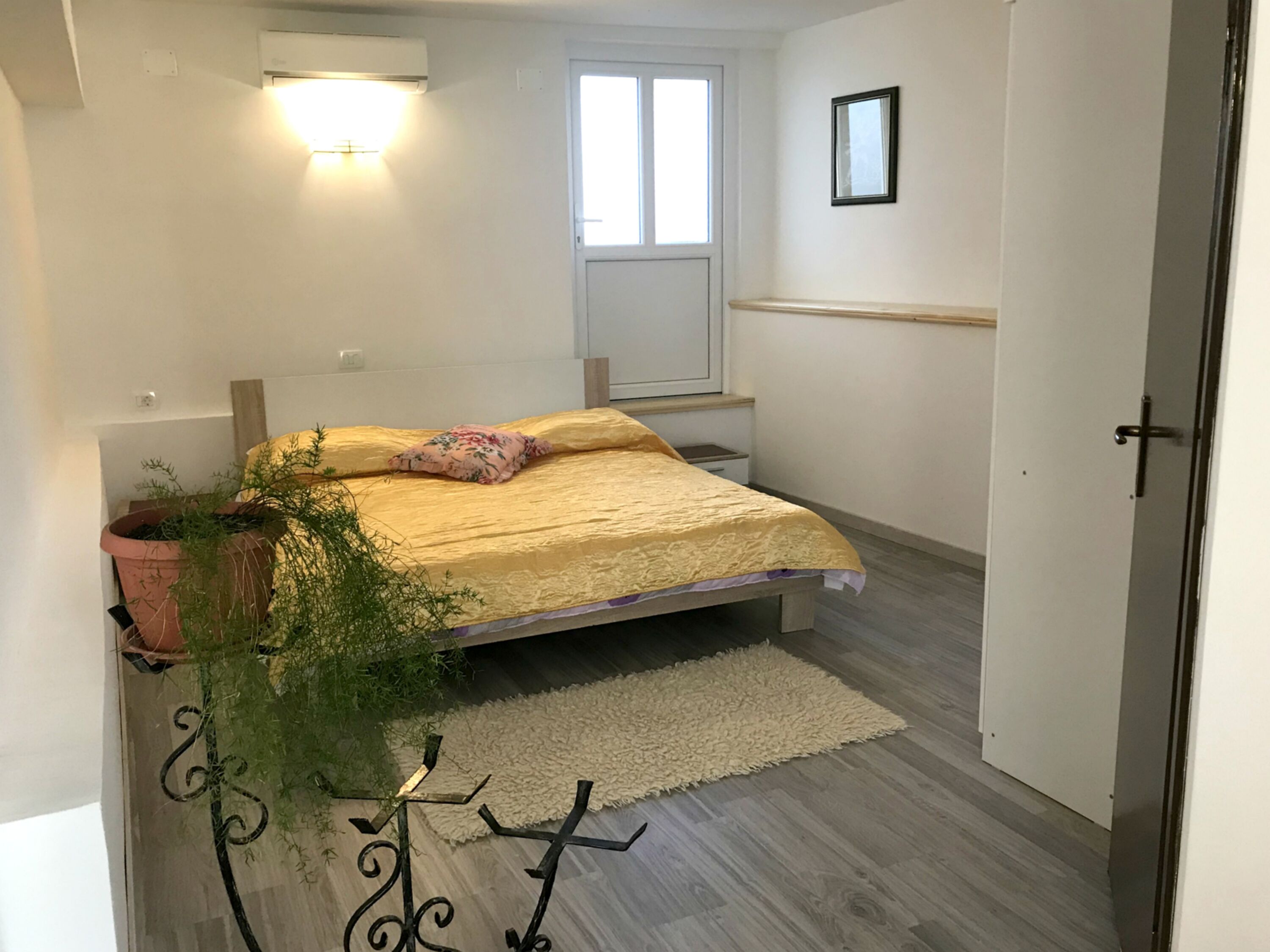 A bedroom with a bed, a potted plant, a window, a door, and a wall-mounted light fixture.