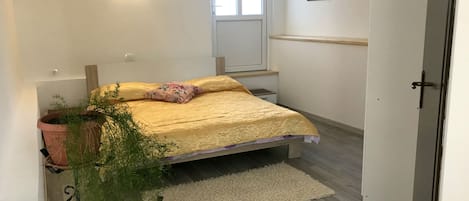 A bedroom with a bed, a potted plant, a window, a door, and a wall-mounted light fixture.
