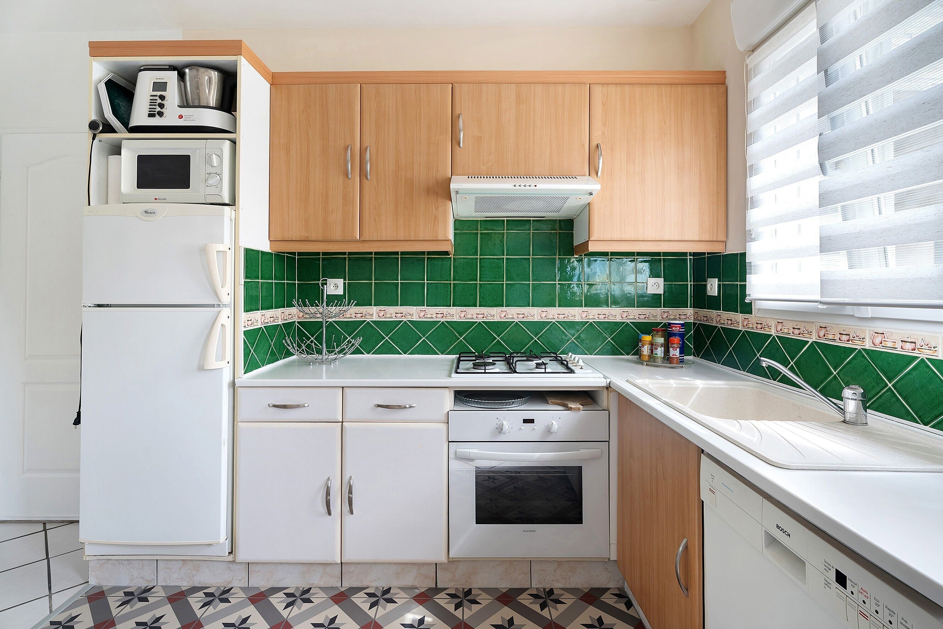 Een compacte keuken met een magnetron, oven en spoelbak, houten kastjes en een groene betegelde achterwand.