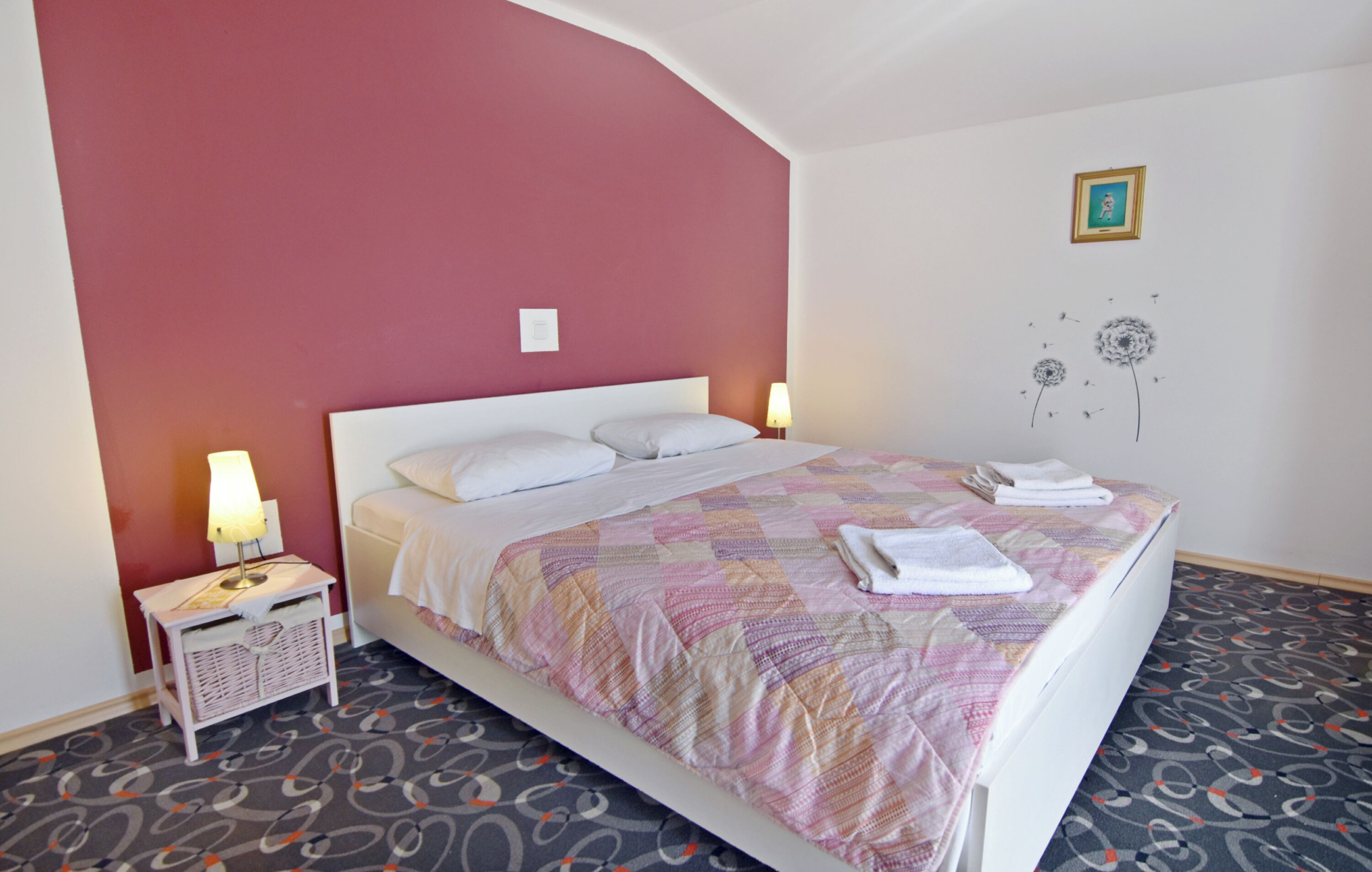 A neatly made bed with a patterned bedspread, two bedside lamps, a small bedside table with a lamp, and a wall with a framed picture and a dandelion illustration.