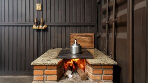 Ein Backofen aus Ziegelsteinen mit einer Metallkessel darauf, ein Holzregal mit zwei Flaschen und ein an der Wand angelehntes Holzbrett.