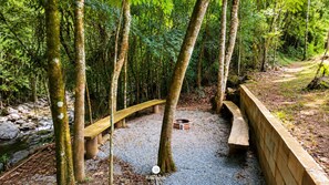 A forested area with a gravel path, wooden benches, and a small fire pit. - Sítio Conteiner do Mano - Águas Mornas (Águas Mornas)