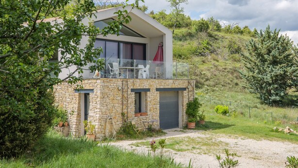 A modern house with a stone base and a glass balcony, surrounded by greenery.