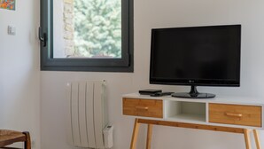 A room with a white desk, a black LG TV, a window with a view of trees, a radiator, and a wooden chair.