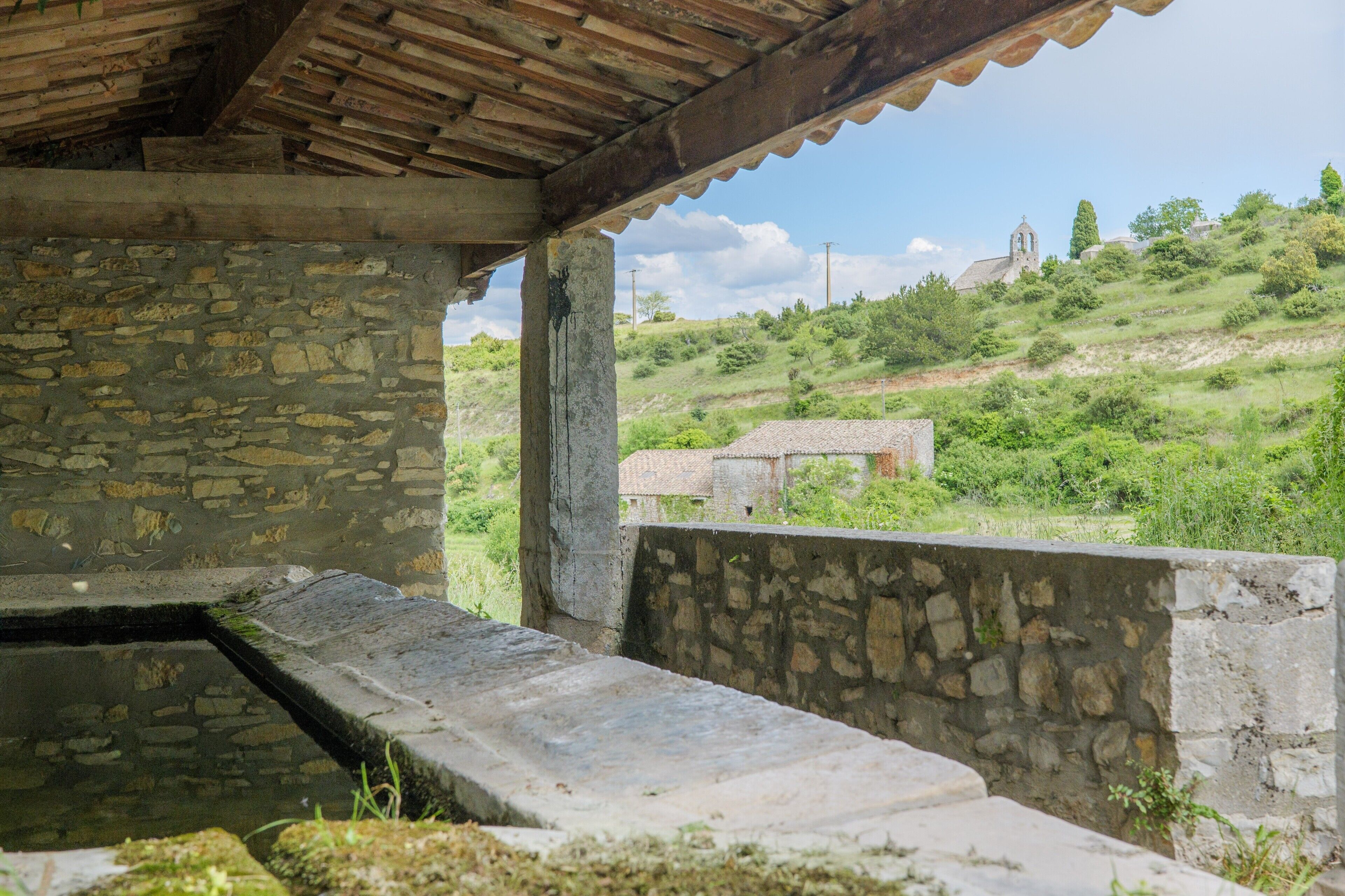 Un edificio de piedra con techo de madera, un muro de piedra y vistas a una colina con una iglesia.