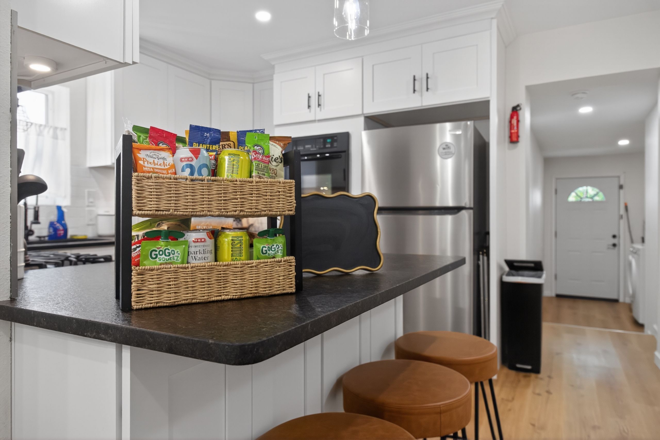 A kitchen with white cabinets, a stainless steel refrigerator, a black microwave, and a wicker basket with snacks on the counter.