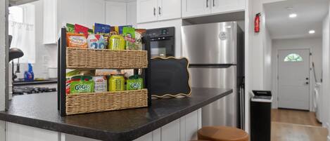 A kitchen with white cabinets, a stainless steel refrigerator, a black microwave, and a wicker basket with snacks on the counter.