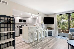 A modern kitchen with white cabinetry, a central island with bar stools, a flat-screen TV mounted on the wall, and a sliding glass door leading to a patio. - Coastal Beach Seascape Villa Closest to Beach with Pools (Hilton Head Island)