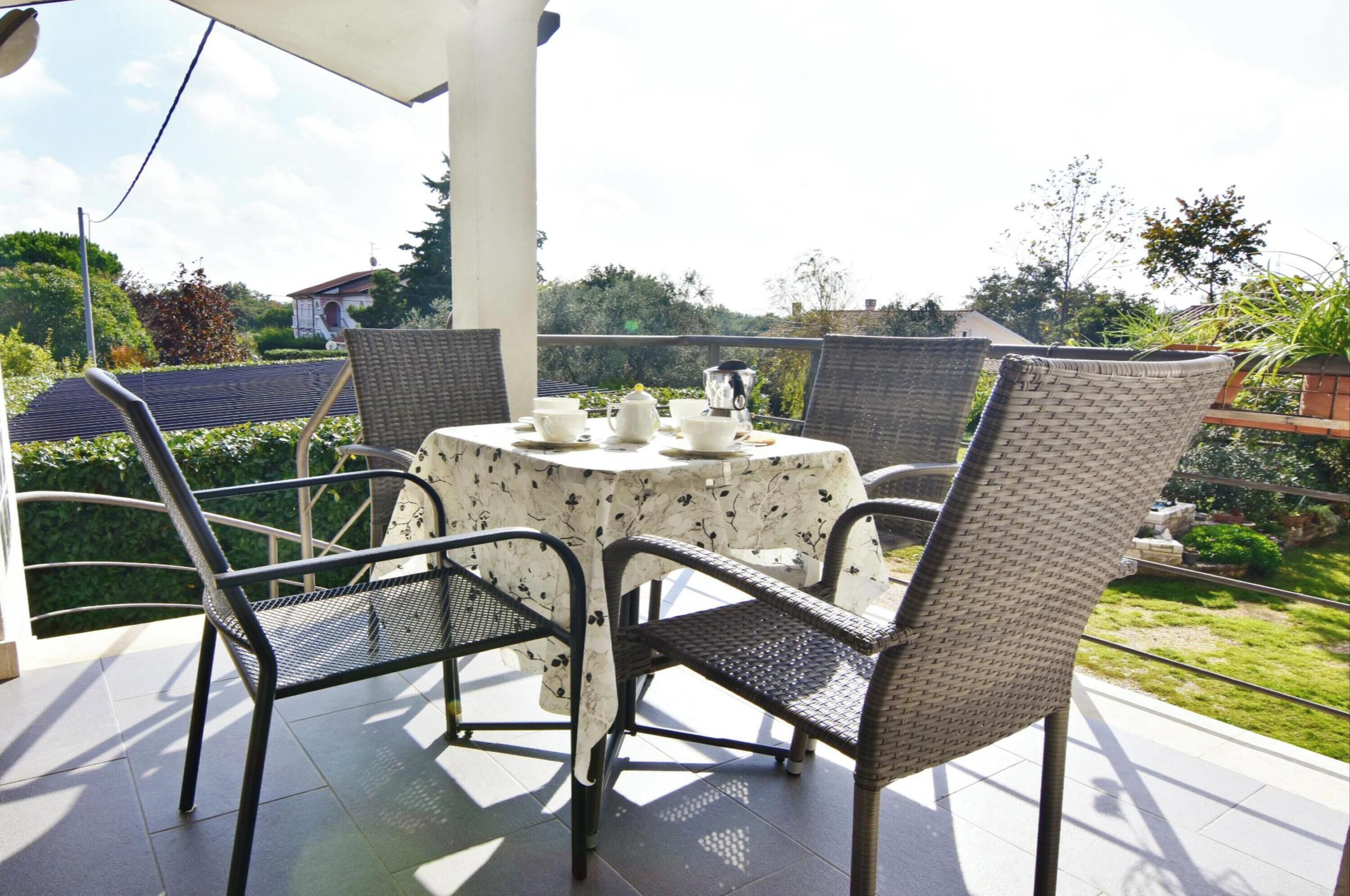 Een overdekt terras met een ronde tafel en vier rieten stoelen, omgeven door begroeiing en uitzicht op een huis.