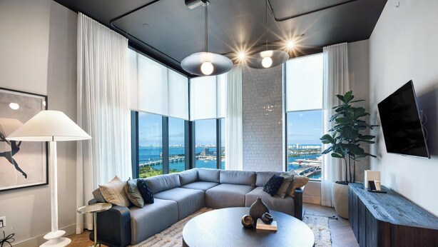 A modern living room with a gray sectional sofa, a round coffee table, and a large window offering a city view. - Bright apartment w/ pool & workspace (Miami)