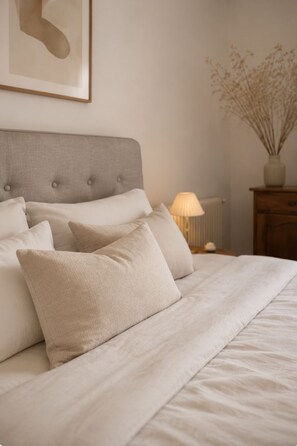 A neatly made bed with a tufted headboard, a bedside lamp, and a vase with dried plants. - Hazel Trees · Porch and Sunset (Saint-Montan)