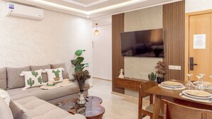 A modern living room with a sofa, coffee table, dining table set for four, and a flat-screen TV mounted on a wooden panel. - Comfortable apartment in the heart of Guéliz (Marrakech)