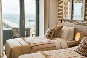A hotel room with a bed, pillows, a bedside table with a lamp, a view of the ocean, and a balcony with chairs. - Hidden 14- Garden Terrace (St Ives)