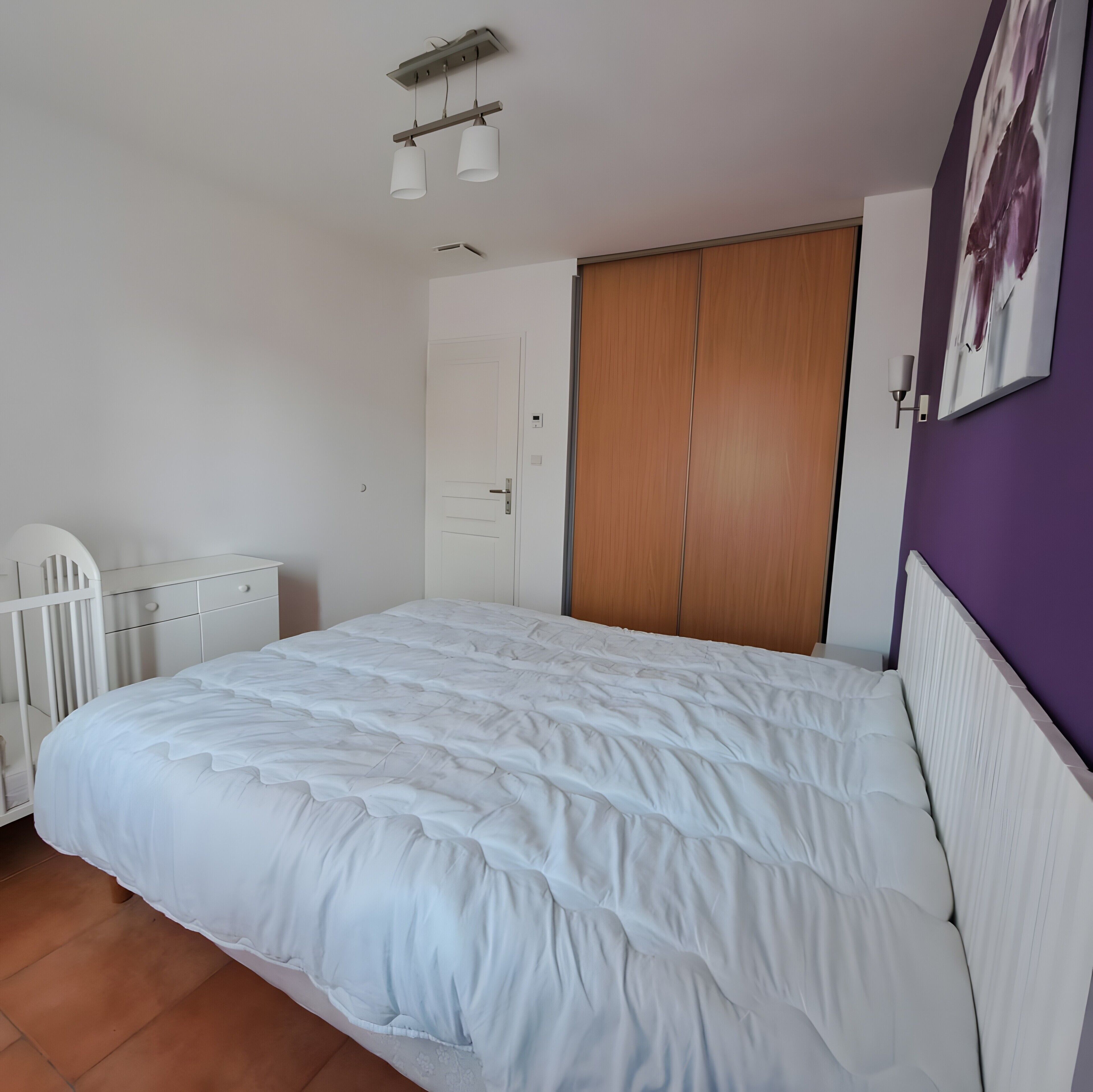 A bedroom with a bed, a wardrobe, a crib, and a ceiling light fixture.