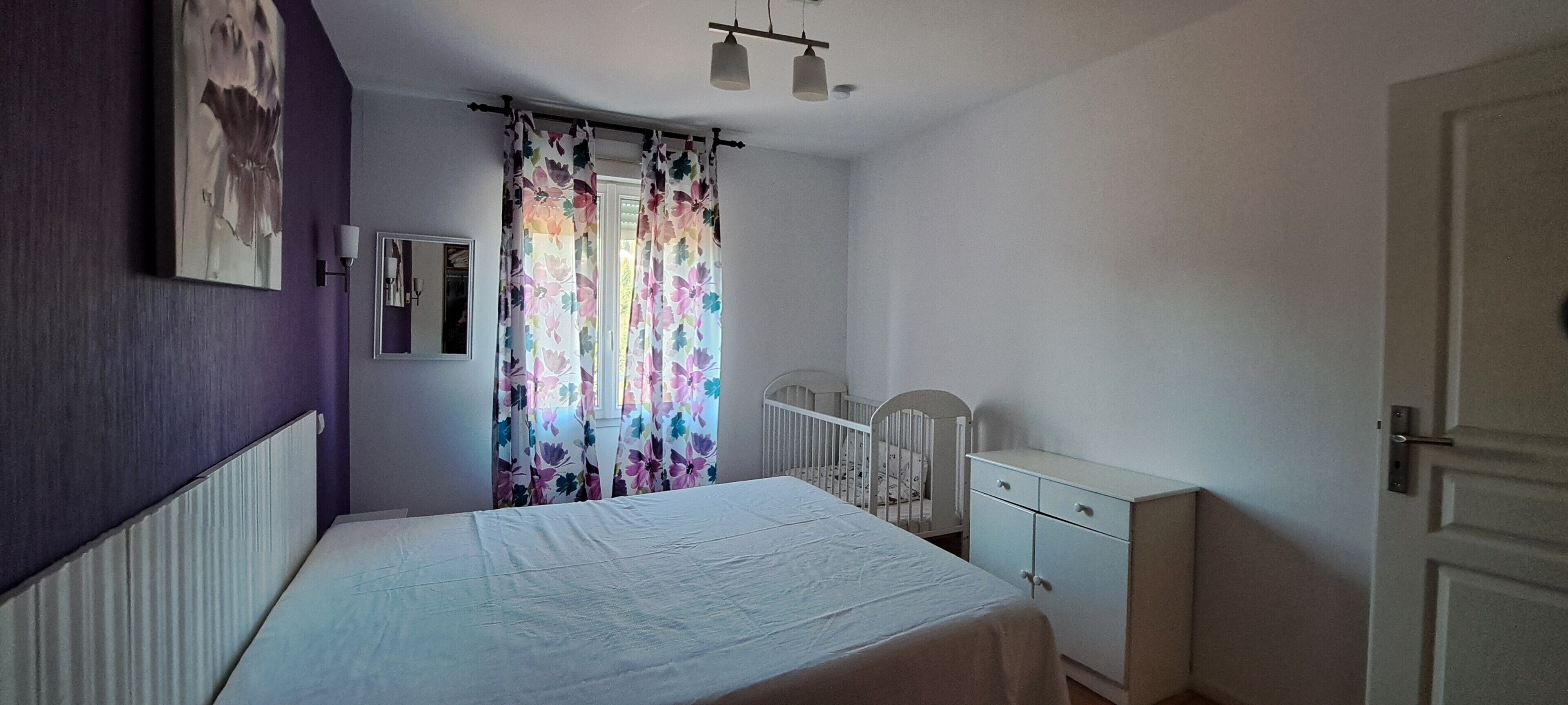 A bedroom with a bed, a crib, a dresser, a wall-mounted lamp, and a window with floral curtains.