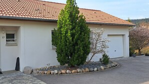 Une maison avec un toit en tuiles, une porte blanche, une petite fenêtre avec un balcon et un jardin bien entretenu, agrémenté d’un conifère et d’un jeune arbre.