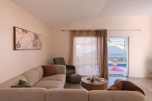 Ein Wohnzimmer mit einer Couch, einem Sessel und einem runden Couchtisch. Eine Glasschiebetür führt zu einem Poolbereich mit einem Flamingoboje.